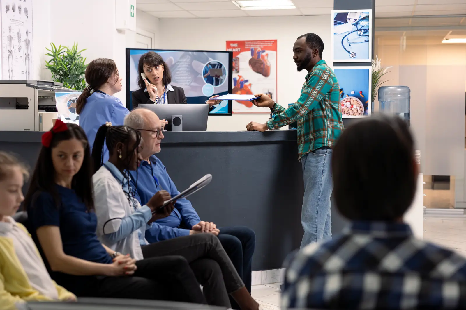 A crowded clinic front desk — a receptionist juggling a phone call and paperwork while a patient waits at the counter